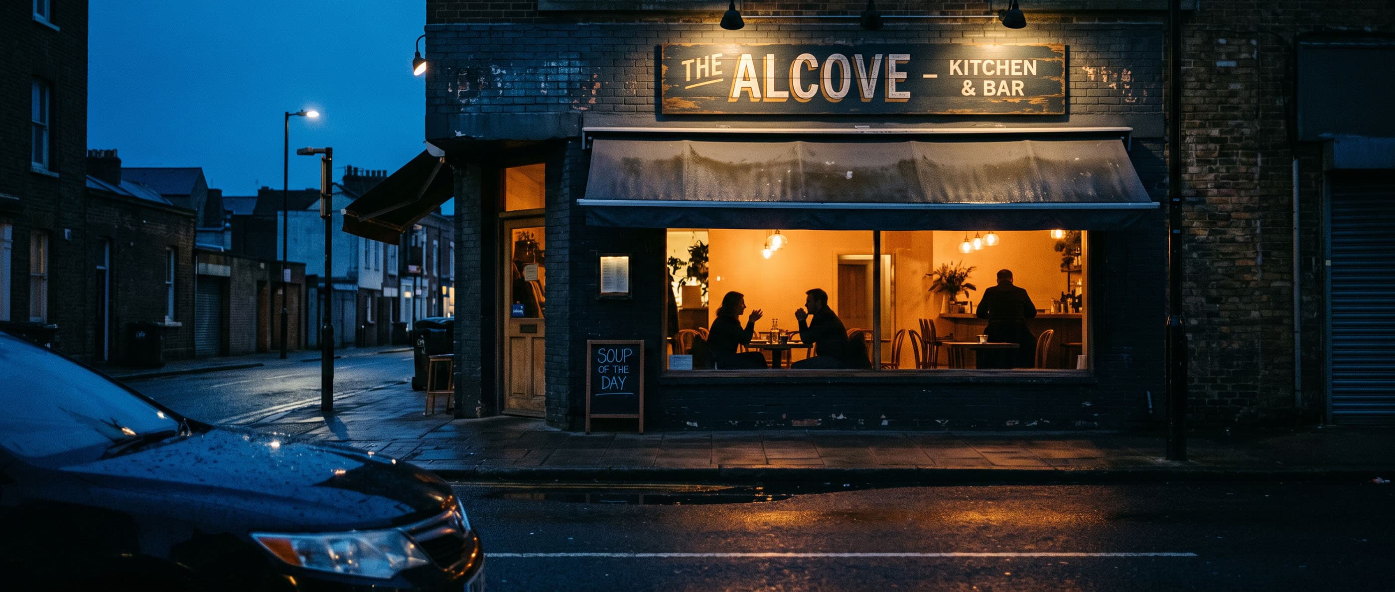 An independent kitchen and bar at dusk, warm light spilling from its windows onto a quiet wet street.